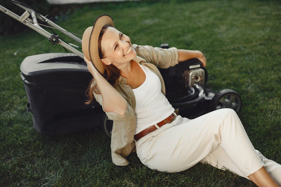 A smiling woman with light skin, long brown hair, and wearing a straw hat, beige shirt, white trousers, and a brown belt, sits on the grass in a garden setting. She is leaning back casually against a 