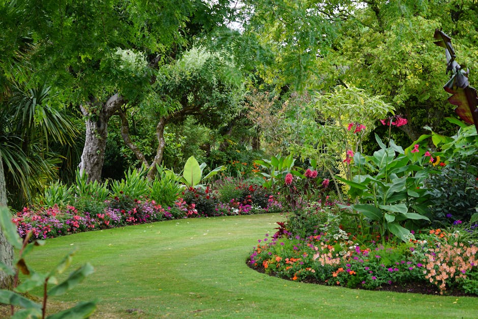 A smiling woman with light skin, long brown hair, and wearing a straw hat, beige shirt, white trousers, and a brown belt, sits on the grass in a garden setting. She is leaning back casually against a 