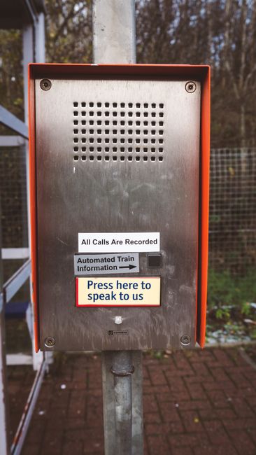 A close-up of an outdoor emergency contact panel mounted on a metal post, featuring a stainless steel surface with small ventilation holes at the top. There is a white label indicating that all calls 